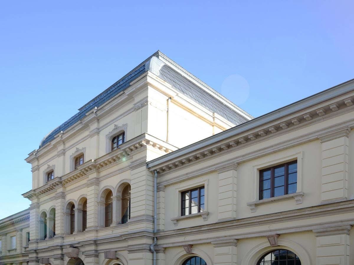 Blick auf das Obergeschoss und den Mittelrisalit des Marstalls in der Marstallstraße. Geputztes hellgelbes Gebäude. Verzierungen aus Sandstein im gleichen Farbton. Die Fenster sind mit Sprossen fast schwarz. Prunkvoller Bau mit Stilelementen des Barock. Blauer Himmel ohne Wolken im Hintergrund.