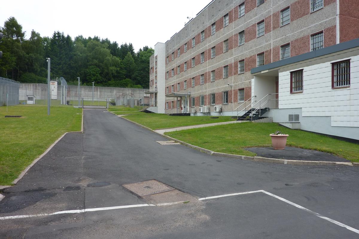 Zu sehen ist ein Gebäude mit vier Etagen. Im Hintergrund befindet sich eine Betonmauer. Davor ist Rasen. Hinter der Mauer sind Bäume zu sehen.