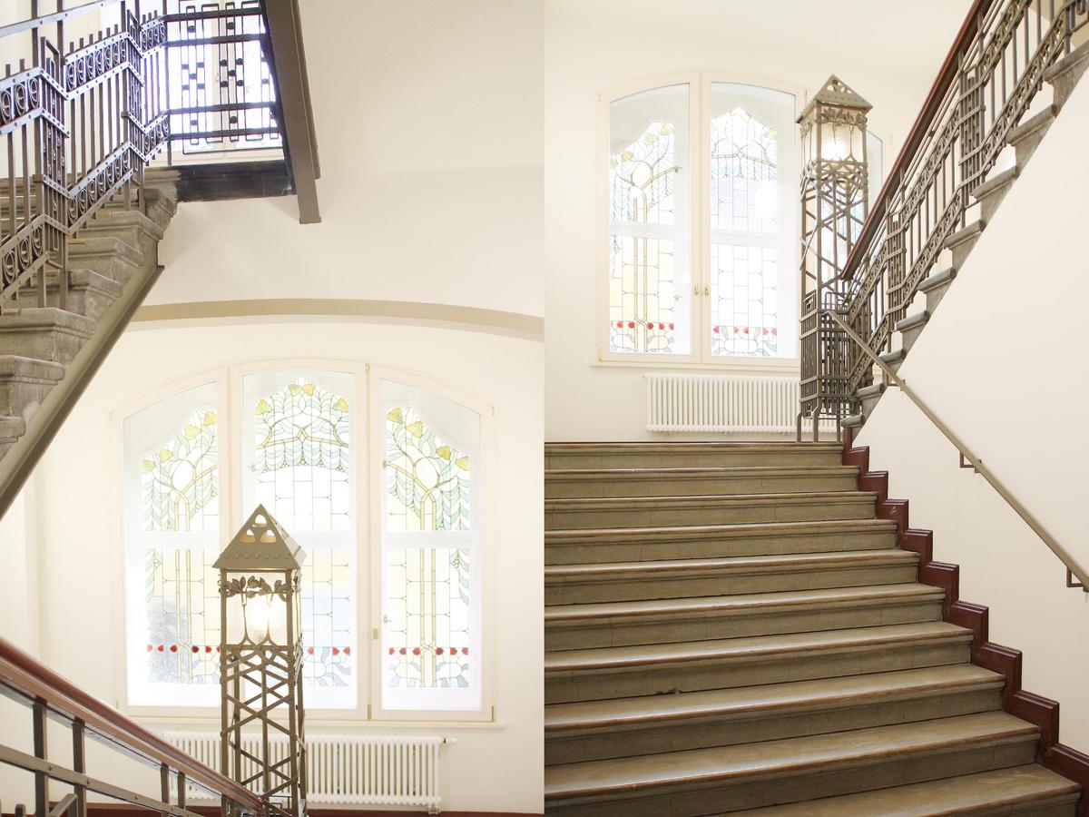 Doppelfoto vom Historischen Treppenhaus, dunkle fast schwarze Treppenstufen, weiße umgebende Wände. Aufwändiges schmiedeeisernes Geländer in dunkelgrau. Rundbogenfenster im Hintergrund mit bunter Bleiverglasung.