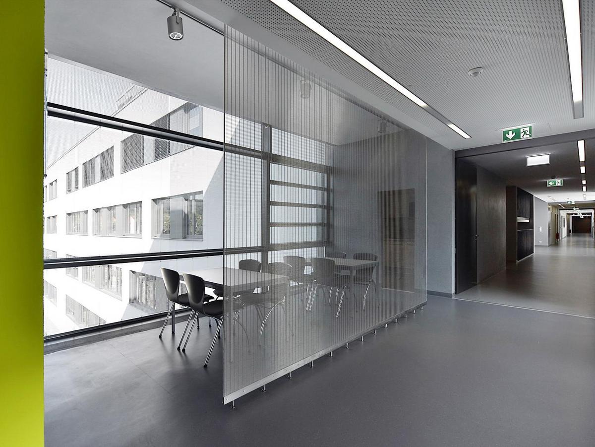 Moderner Innenraum mit Blick in einen dunklen Flur, strichartige lange Lichtleisten an der Decke. Links eine gelbe Wand ist angeschnitten. Ein Gitterrost trennt den Flur vom Aufenthaltsbereich. Dort stehen zwei Tische mit jeweils 4 Stühlen. Außerhalb des Fensters ist wieder die weiße Fassade mit den Fensterbändern zu sehen.