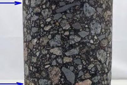 Zu sehen ist ein dunkelgrauer Bohrkern, das heißt eine zylinderförmige Entnahme aus einer Straße. Links sind zwei blaue Pfeile, die auf den Bohrkern weisen.