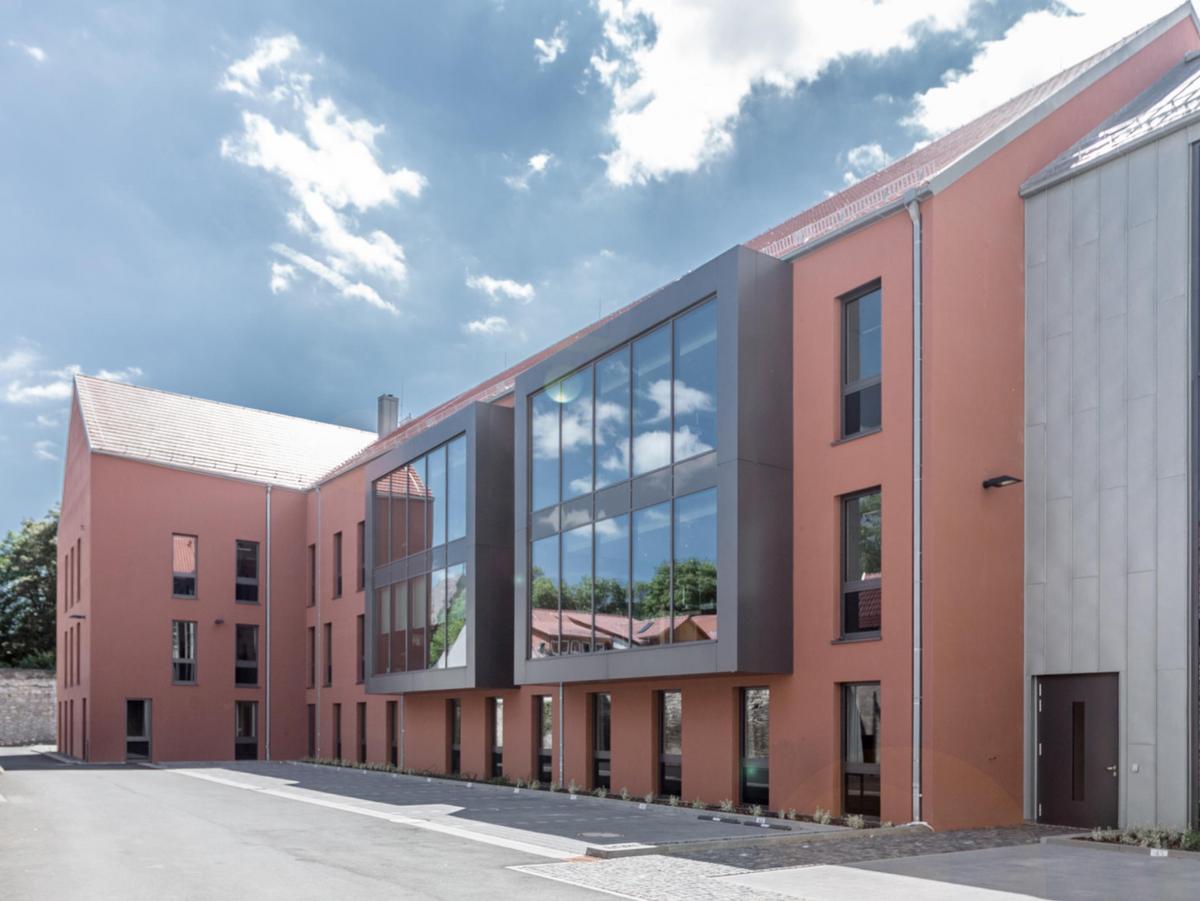 Dreigeschossiger orangener Neubau mit Satteldach. Die Fensterelement sind rasterförmig angeordnet und wirken schwarz. Zwei moderne quadratische graue Erker schieben sich aus der Fassade. Die Dachfläche ist grauen Dachziegeln belegt. Im Vordergrund eine befestigte Fläche und Parkplätze, rechts der Verbindungsbau aus Titanzink und viel Glas. Heiteres Wetter.
