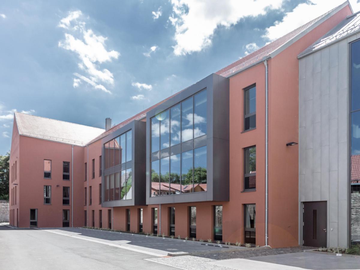 Dreigeschossiger orangener Neubau mit Satteldach. Die Fensterelement sind rasterförmig angeordnet und wirken schwarz. Zwei moderne quadratische graue Erker schieben sich aus der Fassade. Die Dachfläche ist grauen Dachziegeln belegt. Im Vordergrund eine befestigte Fläche und Parkplätze, rechts der Verbindungsbau aus Titanzink und viel Glas. Heiteres Wetter.