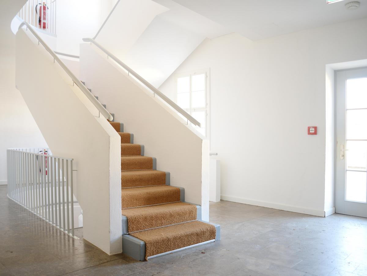 Ein kleines Foyer mit Steintreppe mit Bast-Teppich-Belag und massivem Brüstungsgeländer führt in das Obergeschoss. Rechts kann man das Gebäude verlassen. Der Boden ist hellgrau.