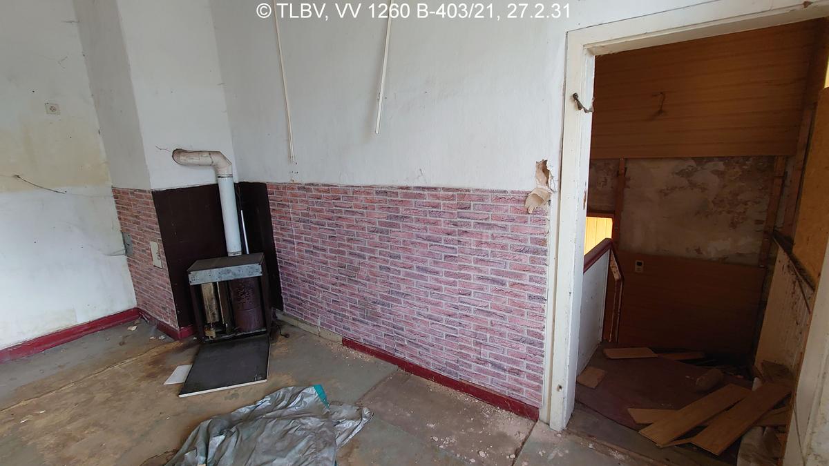 Zimmer mit Ofen, scheinbar defekt, Fußbodenaufbau teilweise zerstört, Wandanstrich und Innentür verschlissen