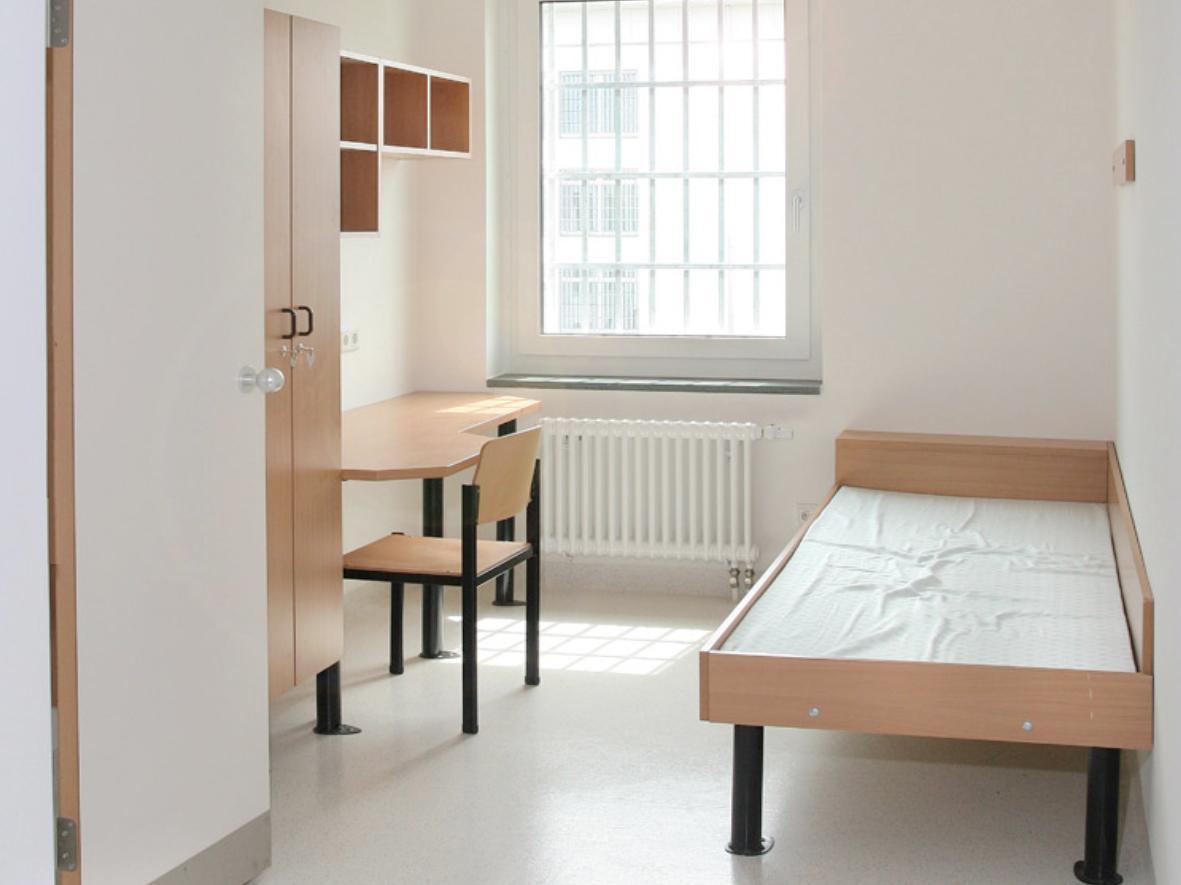 Gefängniszelle: weißer Boden, weiße Wände, weiße Decke mit Fenster mit Vergitterung außen und weißen Heizkörper darunter. Es stehen ein einfaches Bett, ein Tisch mit Stuhl, ein Kleiderschrank und ein Wandregel in L-Form im Raum. Es ist aufgeräumt, steril, kalt.