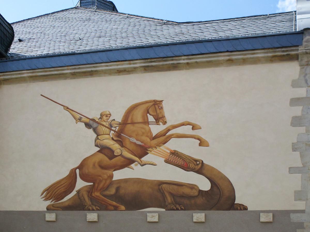Detailbild des Wandgemäldes vom Heiligen Georg an der Fassade im ersten Obergeschoss. Darüber ist das dunkelgraue Dach zu erkennen, welches von der Sonne angestrahlt wird.