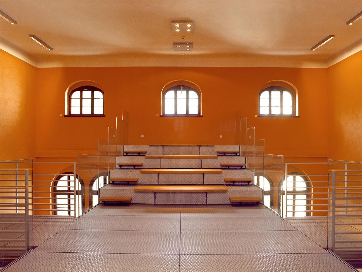 Ein orangener Raum. Im Prinzip eine Brücke die längs das Bild quer. Bodenbelag ist Tränenblech. Brüstungen aus Flachstahl mit Leitereffekt. Man schaut auf Sitzplätze die sich auftürmen und rechts und links davon Treppen um die hinteren Reihen zu erreichen. Hier sind Brüstungen aus Glas, ohne Handlauf. Stirnwand orange mit 3 Rundbogenfenster.