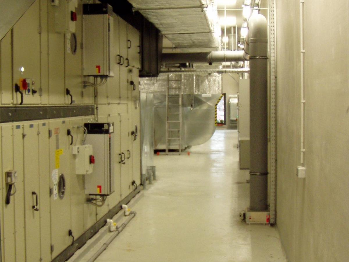 Technikraum in Zentralperspektive, langezogen. An der linken Wand eine Lüftungsanlage, optisch wie ein Schrank, über die komplette Raumhöhe und Raumlänge. Estrichboden, rechts Sichtbetonwand, an der läuft ein rechteckiges Lüftungsrohr in Isolation (silber) eingepackt. An der rechten Wand Kabelinstallationen und eine Kabeltrasse unter der Decke.