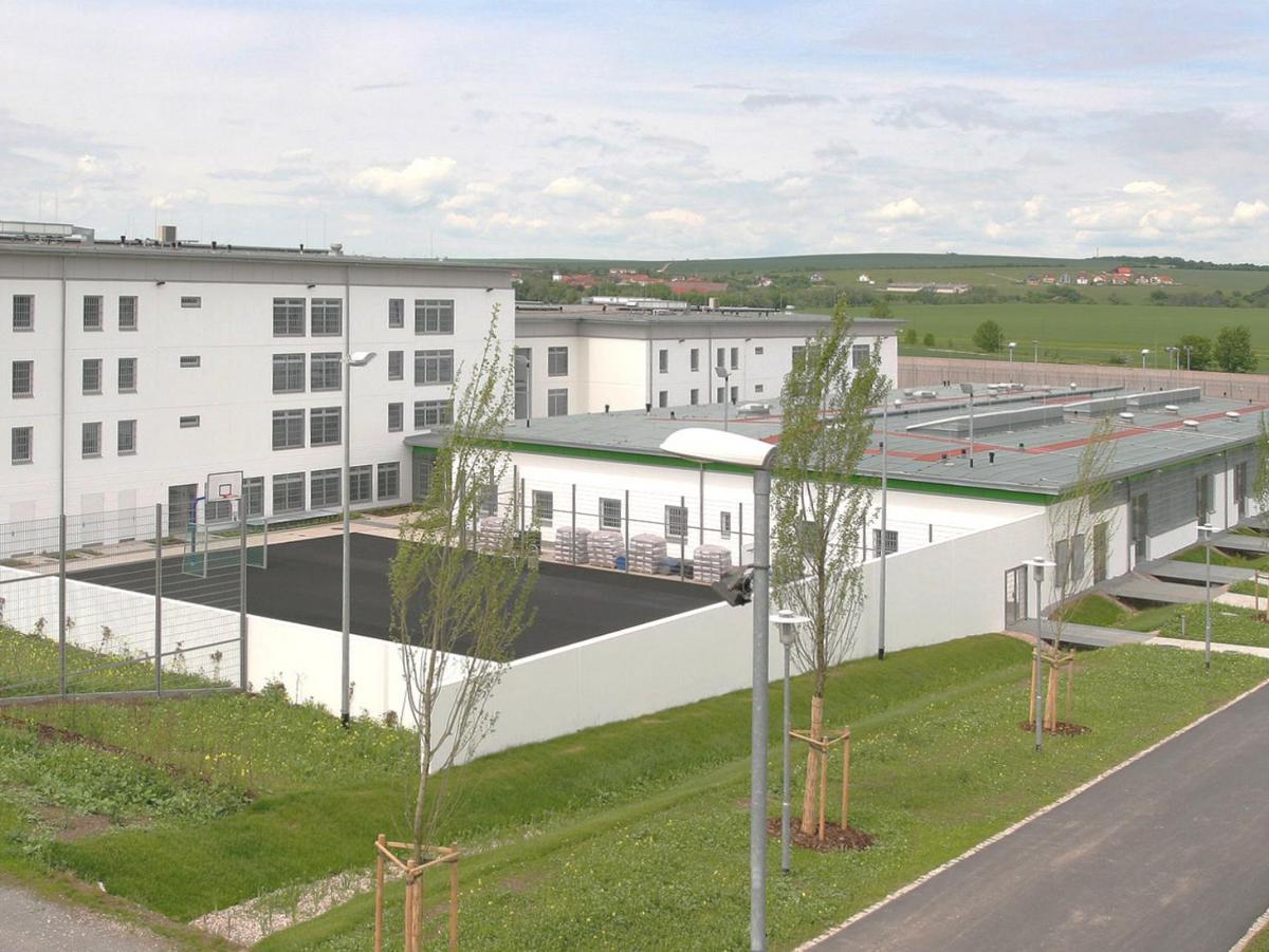 Luftbild mit weißen Mauern im Zentrum und vier- bzw. dreigeschossigen Zellengebäuden dahinter. Im Vordergrund ein Sportplatz aus Teer mit Handballtor und Basketballkorb. Im Vordergrund verläuft ein Versorgungsweg mit jungen Bäumen alleenartig beplanzt. Leichte Bewölkung. In der oberen Hälfte ein weiter Blick in die grünen Felder.